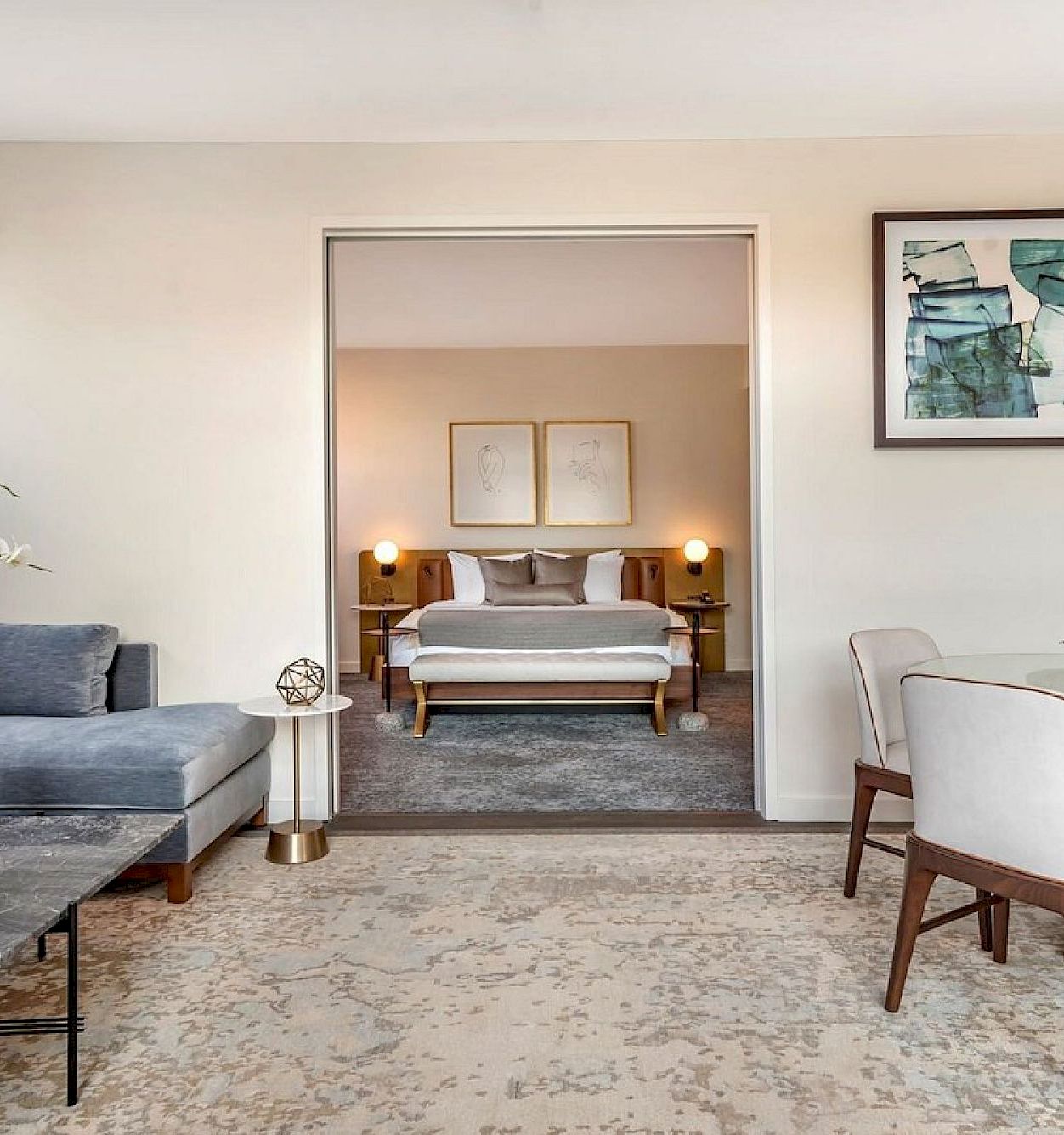 A modern living room and dining area with a sofa, table, flowers, framed art, and an open doorway showing a neatly made bed.
