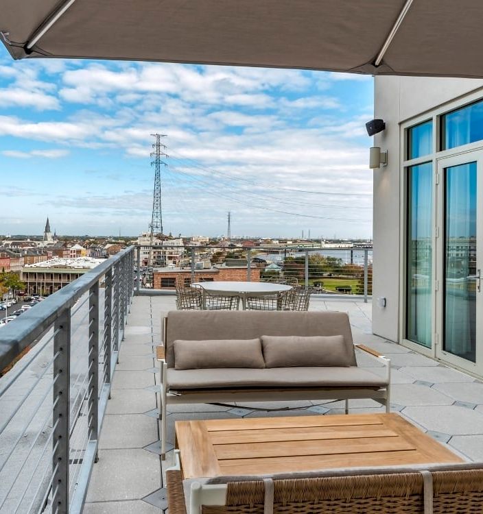 Rooftop terrace with outdoor sofa and table, looking over a city skyline under a blue, cloudy sky.