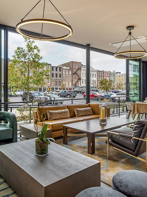 Modern lounge with sofas and dining tables, hanging ring lights, and large windows overlooking a busy street.
