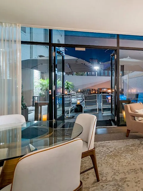 Bright modern living room with sofas, glass doors to a patio dining area, and a large wall-mounted TV behind curtains.