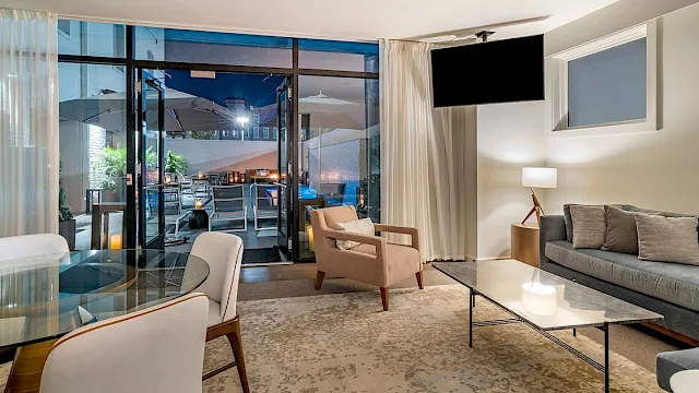 Bright modern living room with sofas, glass doors to a patio dining area, and a large wall-mounted TV behind curtains.