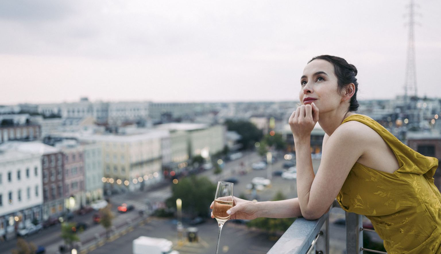 A woman in a yellow dress sips champagne on a rooftop balcony, gazing over a city skyline under an overcast sky.