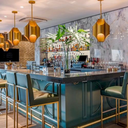 A modern hotel bar with a marble counter, many bottles, blue velvet stools, and gold pendant lights over a back display.