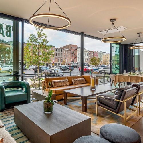Modern lounge dining area with plush seating, tables, and hanging ring lights, overlooking a parking lot and city street.