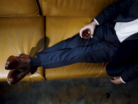 A suited man lounges on a couch, holding a small glass of amber whiskey while his polished leather boots rest nearby.