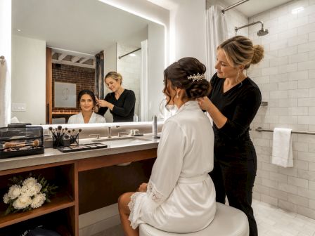 A makeup artist applies makeup to a bride while a second stylist watches in a bright bathroom.
