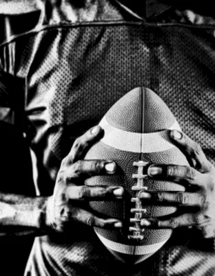 A black-and-white image shows an American football held at chest level by a football player in gear.