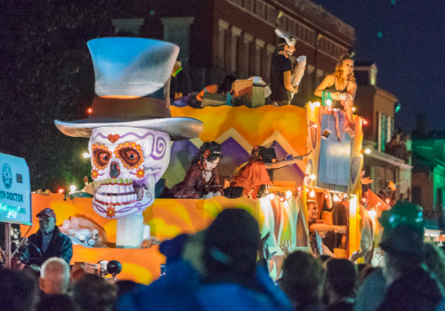 A lively nighttime street parade with giant glowing skull and skeleton floats, musicians, and crowds cheering in colorful lights.