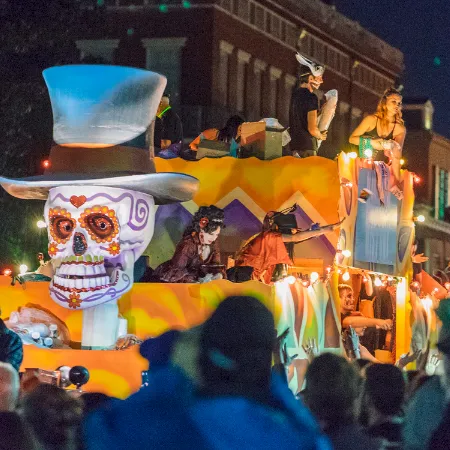 A lively nighttime street parade with giant glowing skull and skeleton floats, musicians, and crowds cheering in colorful lights.