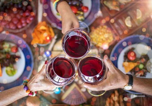 Friends clink glasses of red wine at a festive outdoor table, with colorful drinks and plates in the background, cheers!