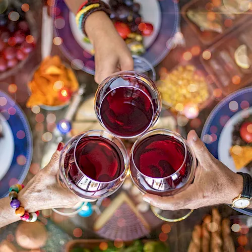 Friends clink glasses of red wine at a festive outdoor table, with colorful drinks and plates in the background, cheers!