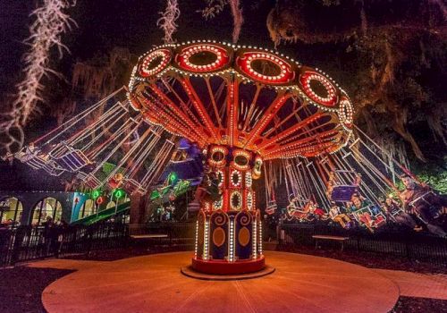A glowing carnival ride spins at night, with a large red-and-gold decorated center and colorful lights around the drum-shaped base.