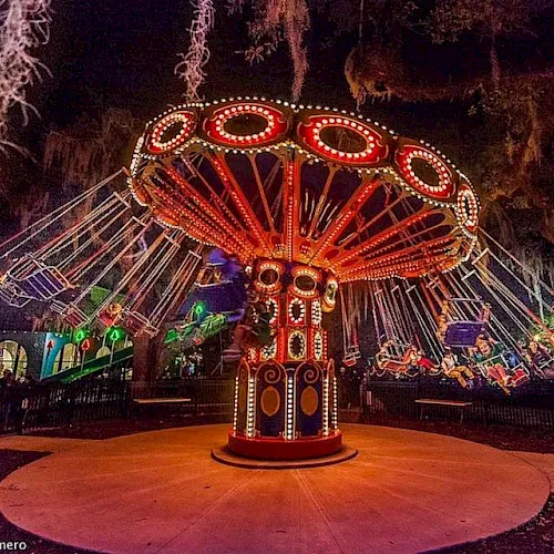 A glowing carnival ride spins at night, with a large red-and-gold decorated center and colorful lights around the drum-shaped base.