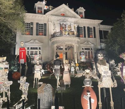 A large white house decorated with Halloween decorations including skeletons and tombstones at nighttime.