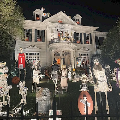 A spooky, Halloween-style haunted house at night, filled with skeleton figures, tombstones, and creepy props on the lawn.