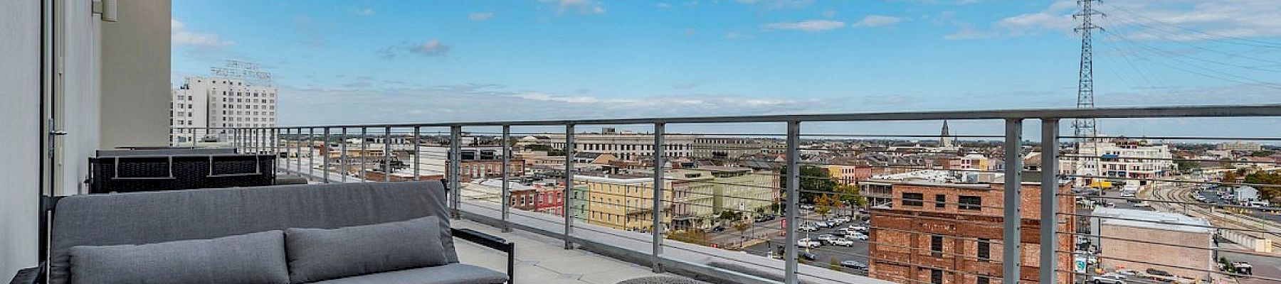 A modern balcony with outdoor seating and a table, overlooking a city skyline under a cloudy blue sky.