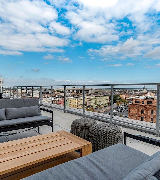 A modern balcony with outdoor seating and a table, overlooking a city skyline under a cloudy blue sky.