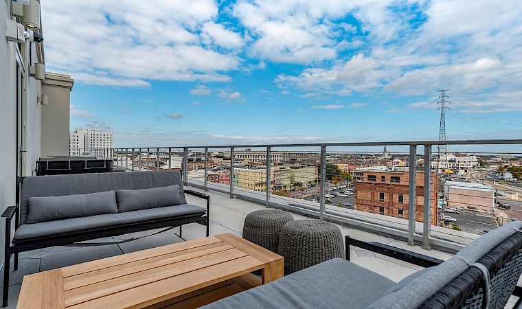 A modern balcony with outdoor seating and a table, overlooking a city skyline under a cloudy blue sky.