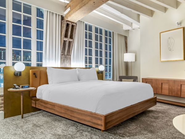 A modern hotel bedroom with a large bed, warm wood headboard, floor lamp, curtains, and city-view windows at night.