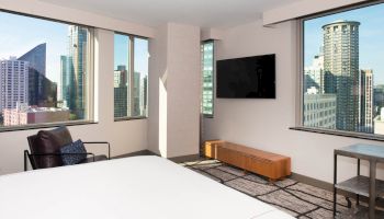 A modern hotel room with large windows showcasing a city view, a TV on the wall, a bed, and a chair. Ends the sentence.