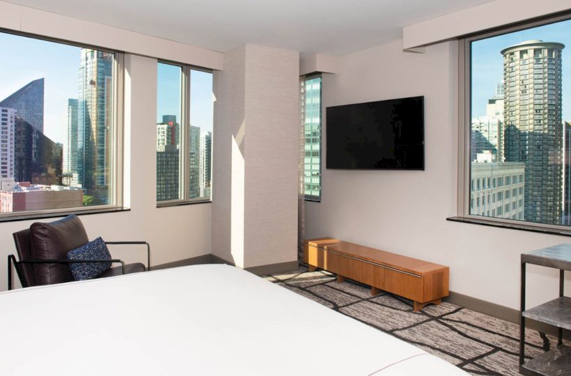 A modern hotel room with large windows showcasing a city view, a TV on the wall, a bed, and a chair. Ends the sentence.