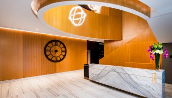 A modern lobby features a marble reception desk, a large wall clock, wooden accents, stylish lighting fixtures, and a vase with colorful flowers.