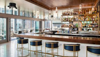 The image shows a modern bar with a well-stocked back shelf, high-top chairs, hanging lights, and large windows offering a city view.