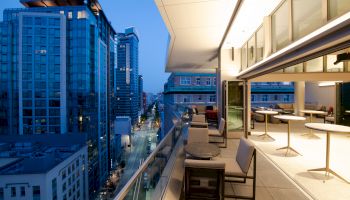 A modern cityscape in the evening from a terrace featuring tables and chairs, overlooking high-rise buildings and a street below.