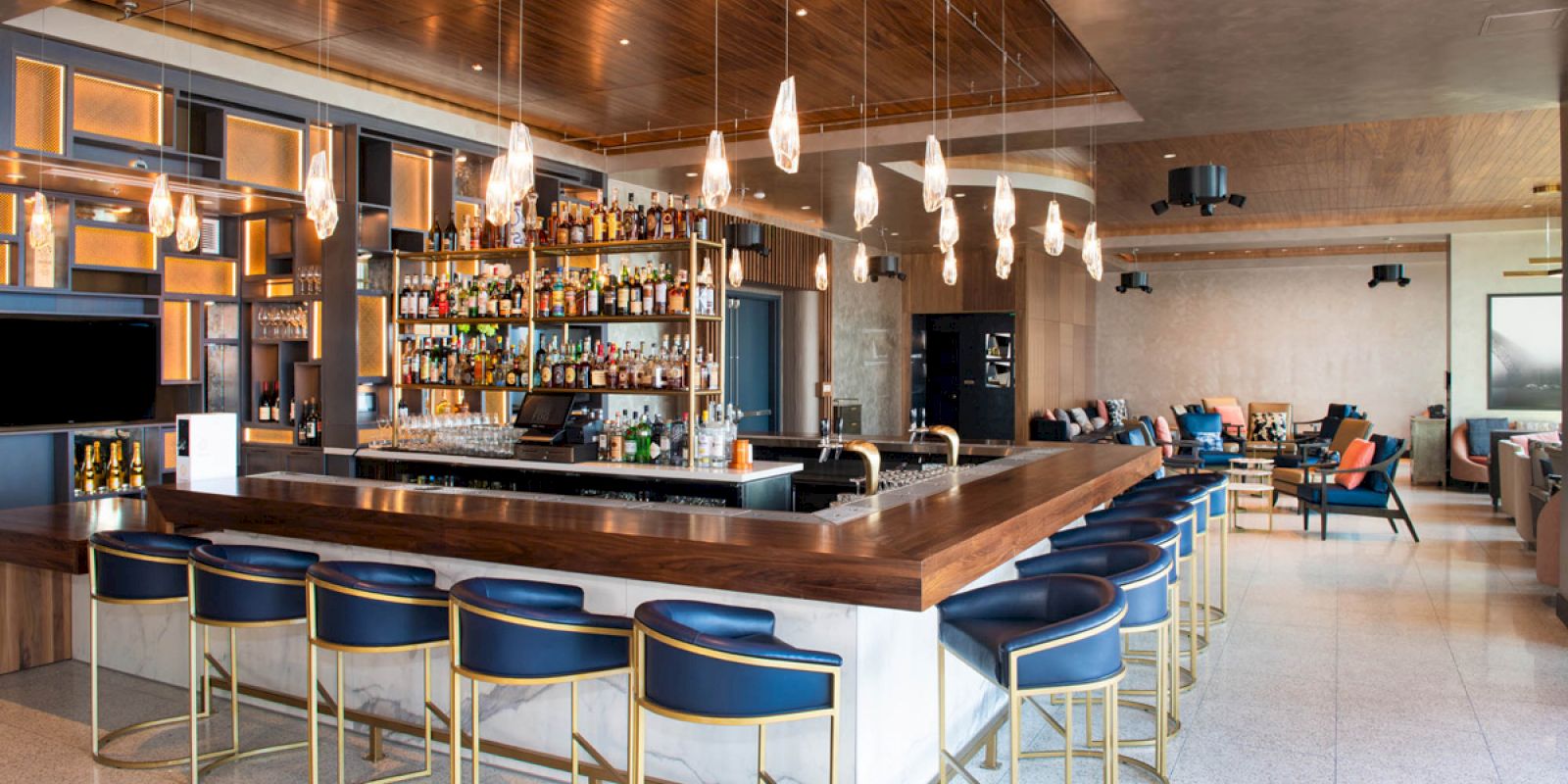 This image shows a modern bar with a well-stocked liquor shelf, stylish seating, pendant lights, and a relaxed lounge area in the background.