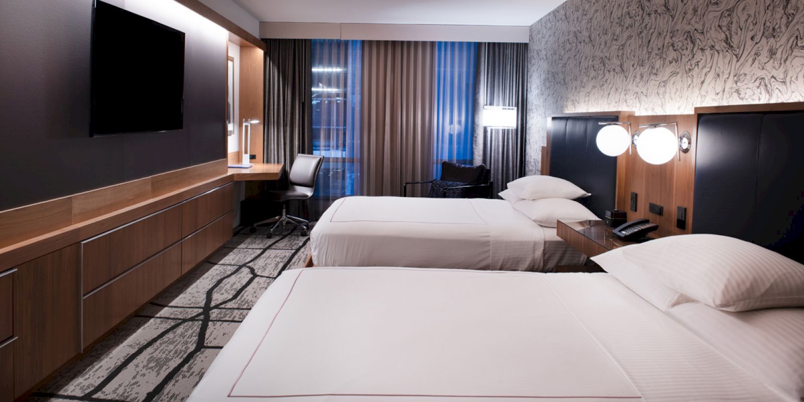 The image shows a modern hotel room with two double beds, a flat-screen TV, a work desk with a chair, curtains, decorative lighting, and a patterned carpet.