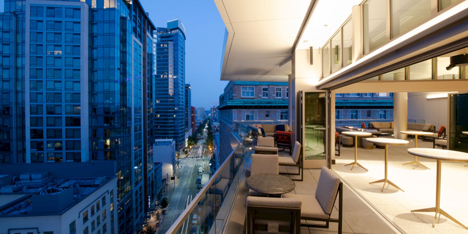 A modern cityscape at dusk featuring tall buildings and a stylish balcony with tables and chairs, offering an elevated view of the street below.