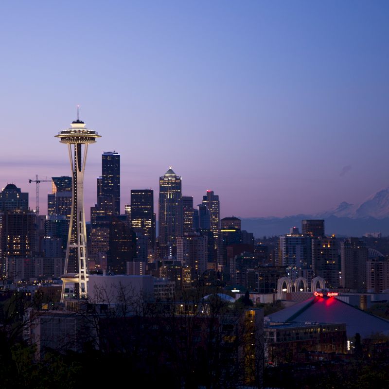 The image shows a city skyline at dusk with the Space Needle prominently featured and a mountain in the background.