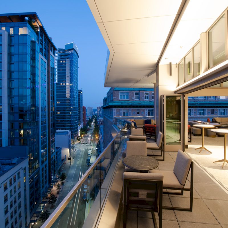 The image shows a modern cityscape at dusk, viewed from a spacious balcony with seating and tables, overlooking tall buildings and a street below.