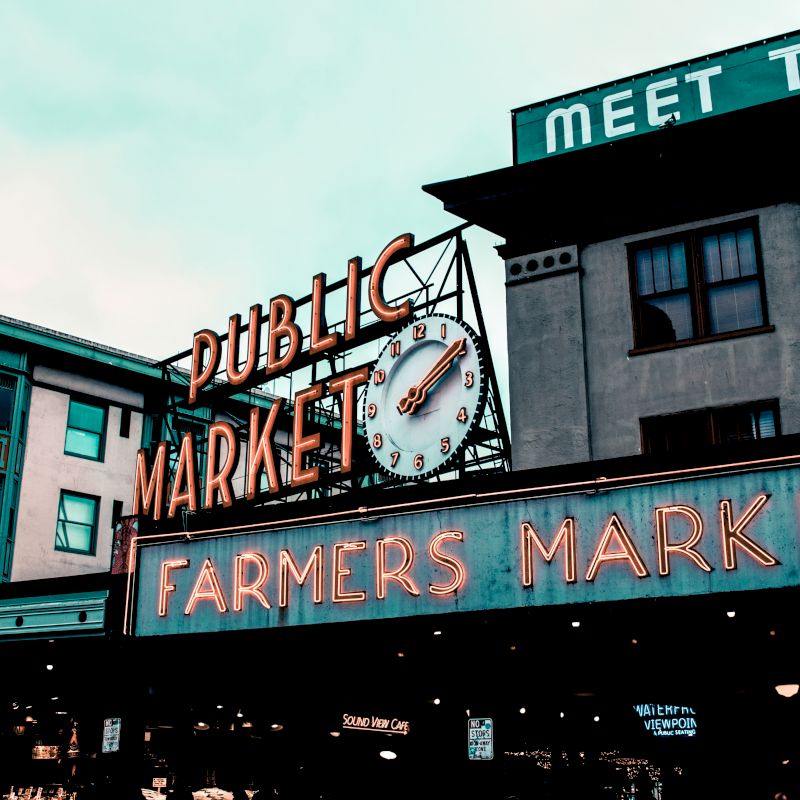 This image shows a neon sign for a public market and farmers market, with buildings in the background. The sign includes a clock displaying the time.