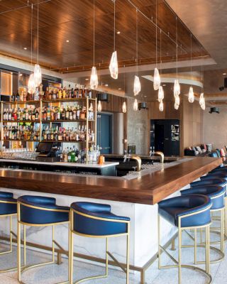 A modern bar with a wooden counter, blue bar stools, hanging lights, and a well-stocked liquor shelf; seating area in the background.