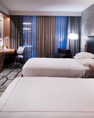 The image shows a modern hotel room with two double beds, a wall-mounted TV, a desk with a chair, and contemporary decor.