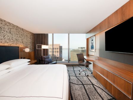 The image shows a modern hotel room with a large bed, a desk, a chair, a TV, and a window offering a cityscape and waterfront view.