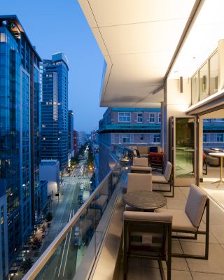 A modern rooftop terrace with seating overlooks a city street lined with high-rise buildings at dusk, under a clear evening sky.