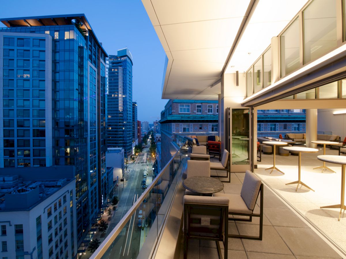 A modern cityscape with tall buildings at dusk, featuring a spacious outdoor balcony with chairs and tables overlooking the street below.