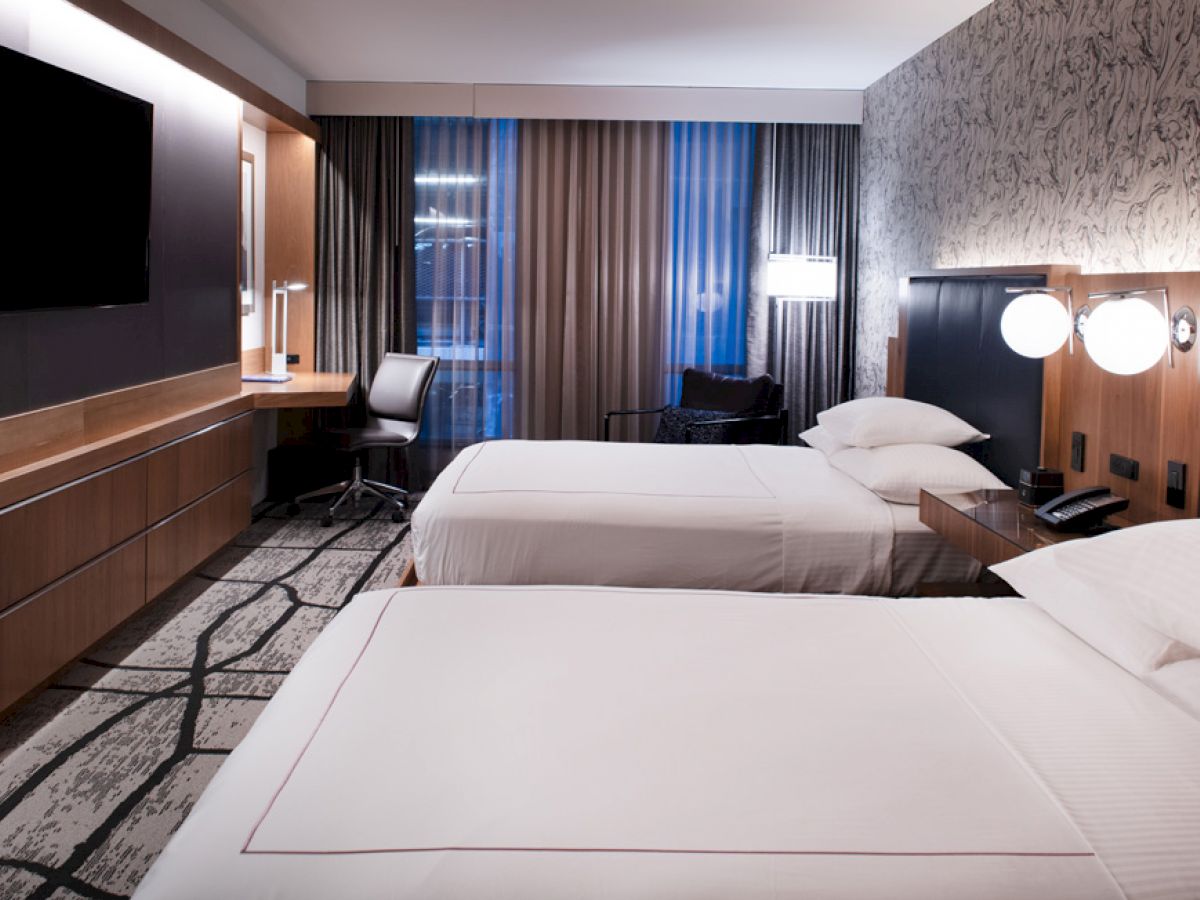 A modern hotel room with two double beds, a wall-mounted TV, a work desk near the window, and stylish decor.