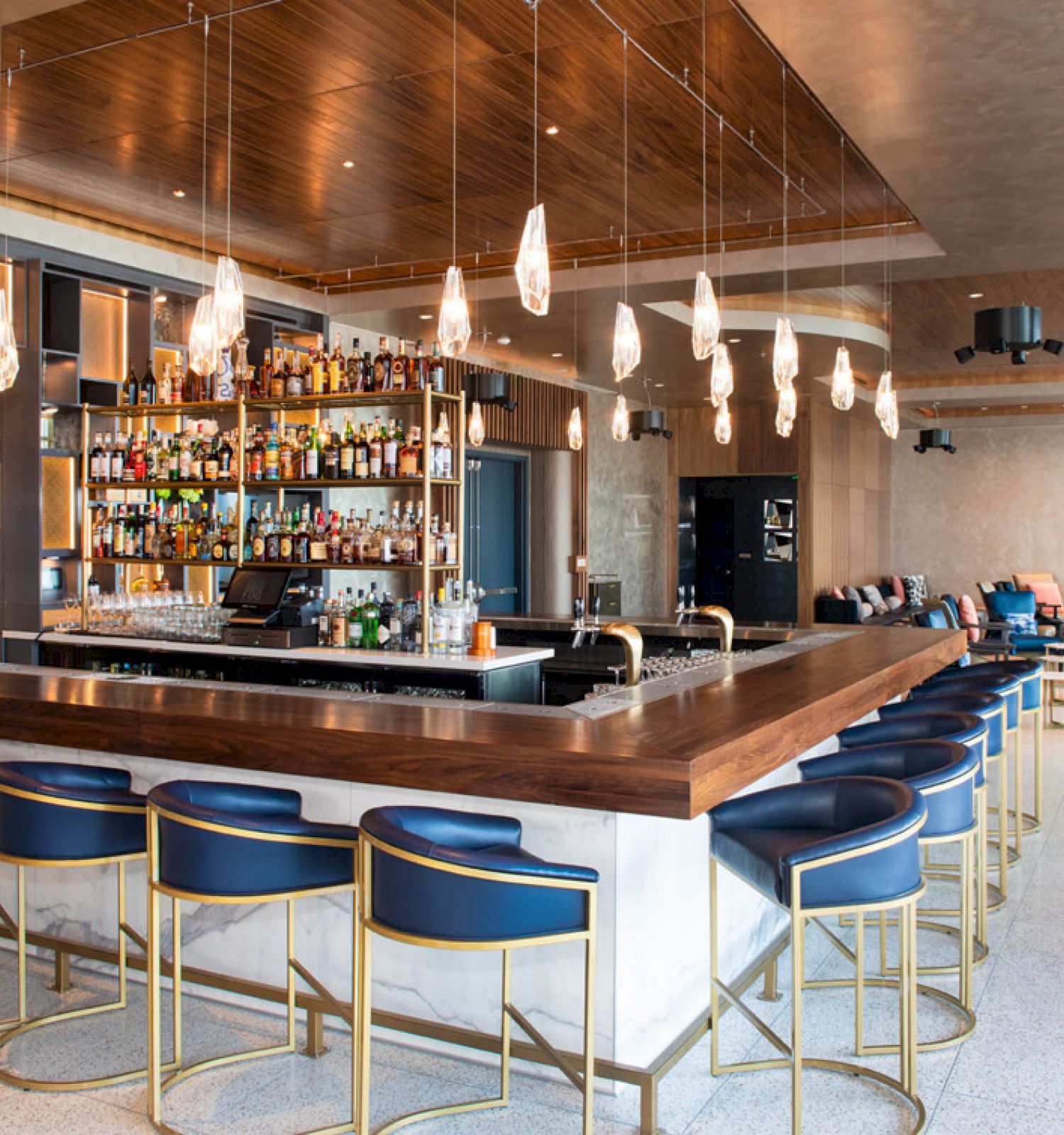 This image shows a modern bar with a long wooden counter, bar stools, overhead lighting, and a well-stocked shelf of bottles in the background.