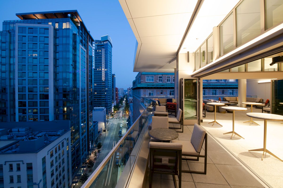 A modern urban balcony with outdoor seating overlooks a city street lined with tall buildings during twilight, capturing a vibrant yet serene atmosphere.