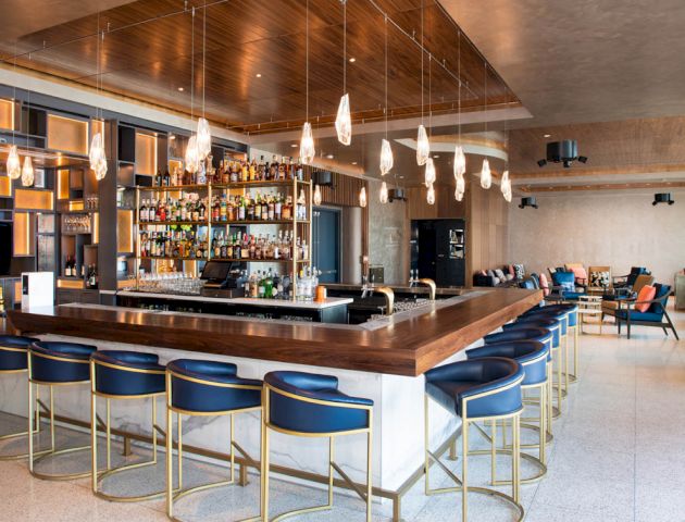 The image shows a modern bar with a sleek wooden counter, blue bar stools, pendant lighting, and a well-stocked liquor shelf.