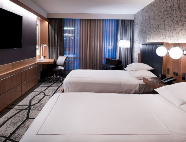 A modern hotel room with two neatly made beds, a flat-screen TV, a desk with a chair, and floor-to-ceiling curtains.