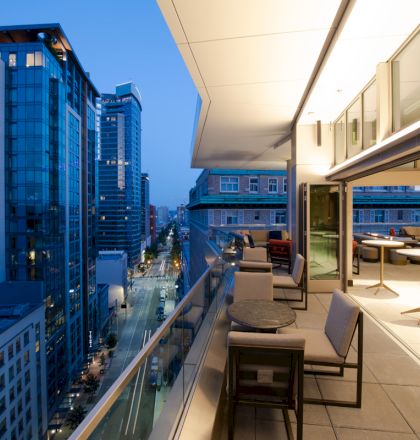 This image shows a modern cityscape at dusk with tall buildings and an outdoor balcony area equipped with seating and tables.