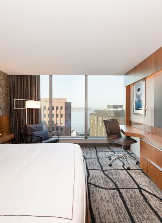 A modern hotel room features a large bed, flat-screen TV, desk, chair, and a window with a city view. The design is sleek and contemporary.