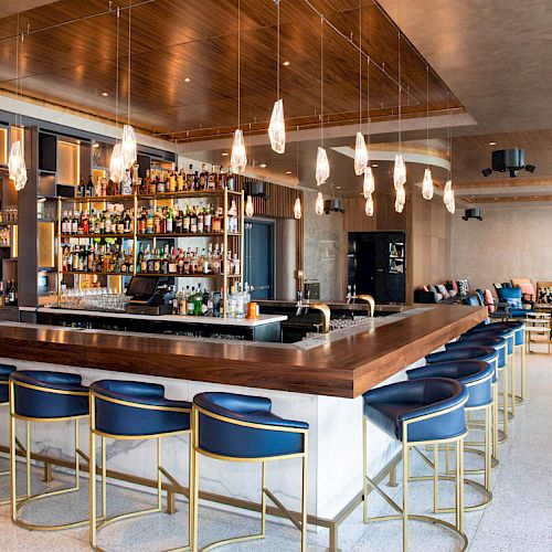 A modern bar with pendant lights, blue cushioned stools, and a fully stocked bar counter in a sleek, contemporary setting.