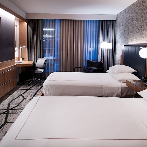 A modern hotel room with two beds, a flat-screen TV, a desk, and large windows with curtains letting in natural light, featuring a chic design.