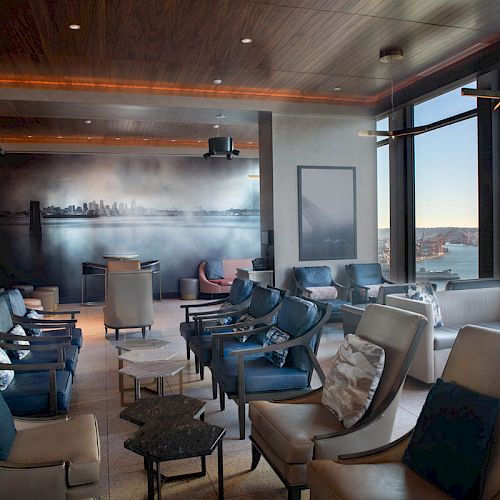 A modern lounge with ample seating, overlooking a scenic waterfront city view through large windows. The room features stylish decor and a mural.