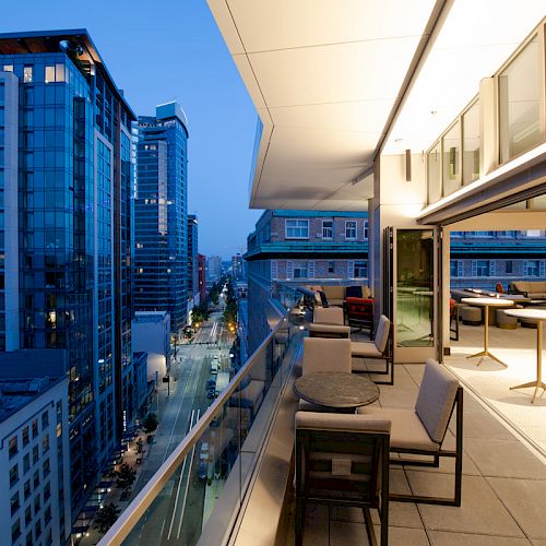 An outdoor balcony in a modern city at dusk, featuring several tables and chairs, with tall buildings and a city street visible below.